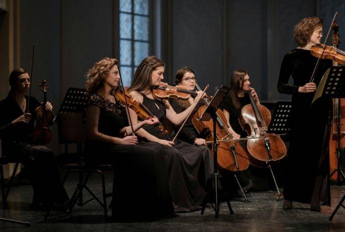 Orchestra with string instruments, players in black attire, performing on stage with focused expressions and formal setting.