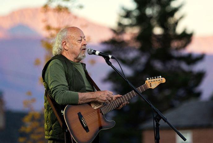 An artist playing an electric guitar and singing into a microphone outdoors with mountains and trees in the background.