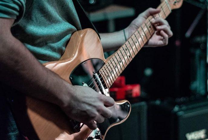 A person playing a light brown electric guitar, focusing on the hands and guitar body closely.