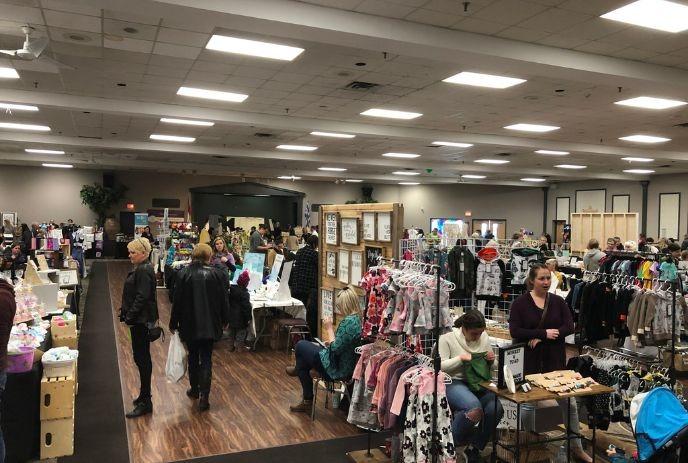 A busy indoor market with various stalls selling clothes, crafts, and other items. People are browsing and shopping.