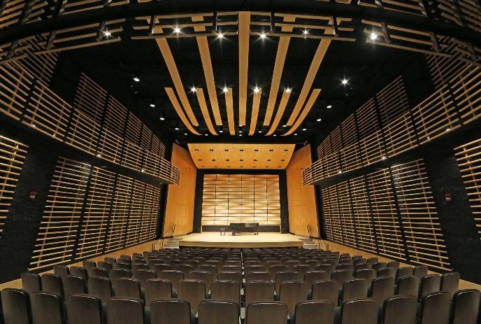 A modern concert hall with wooden paneling, curved slats, rows of chairs, and a stage with a grand piano.