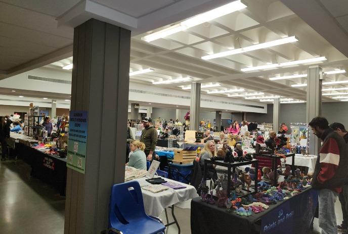 A bustling indoor market with various vendors displaying crafts and goods on tables, under bright ceiling lights.