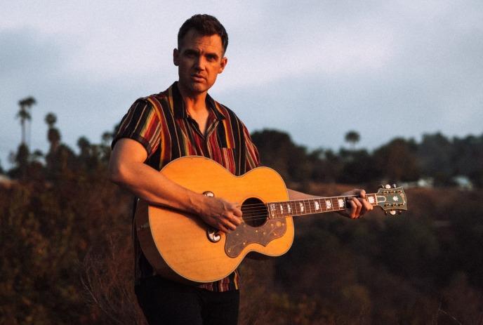 Tyler Hilton holding a guitar outdoors at dusk.