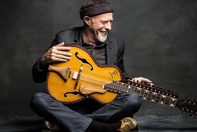 Harry Manx in a black suit playing a yellow guitar while sitting on the ground under