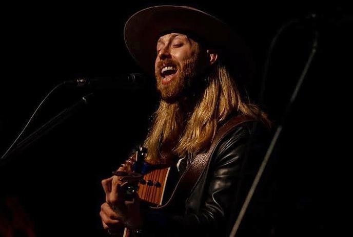 wearing a hat and a leather jacket, is playing an acoustic guitar on stage with dark background lighting.