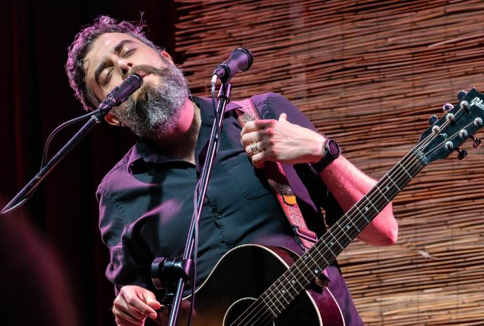 A person playing an acoustic guitar on stage. He wearing a black shirt and a wristwatch, adjusting a microphone.