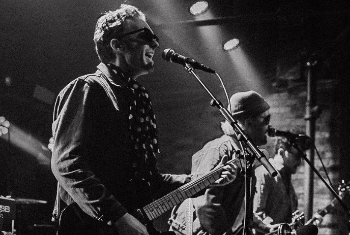 A black and white photo of three musicians performing on stage with guitars and microphones under stage lights.