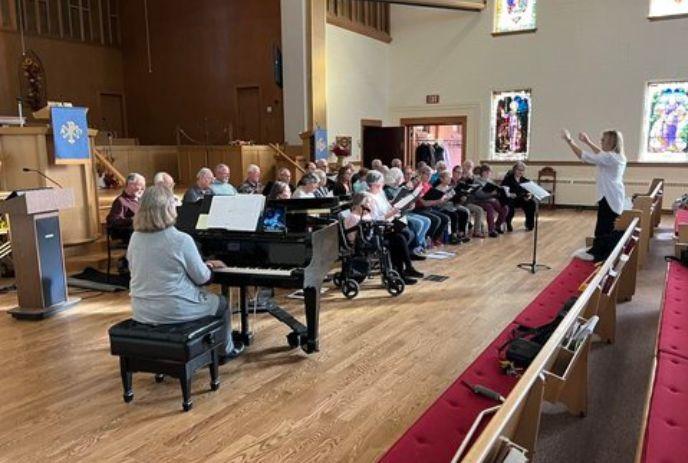 A group of people seated in church-like setting participating in choir practice. One plays the piano while another conducts.