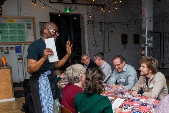 A server holding a piece of paper speaks to a group of people seated around a table in a festive restaurant.