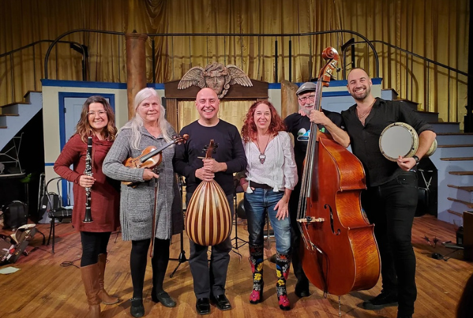 A group of seven musicians on stage with instruments like guitar, oud, double bass, and banjo smiling.