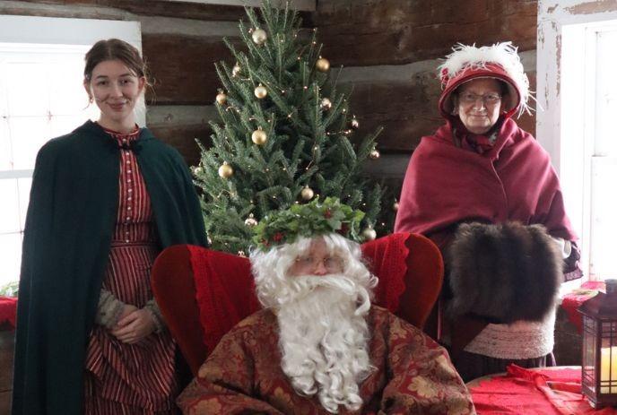 Three in historical attire stand by a decorated Christmas tree: Santa in red chair, others in cloaks and bonnets.