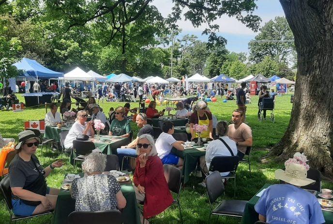 Outdoor festival scene: people at tables under trees, tents and booths in the background, enjoying food and chatter.