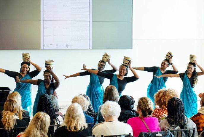 Dancers in blue dresses and black tops perform on stage, balancing baskets on their heads, before an audience.