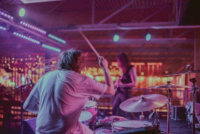 A drummer performing with his drum set and a guitarist in the background under colourful purple and yellow lights