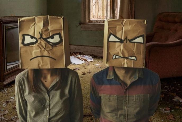 Two people stand in a dilapidated room, each wearing a paper bag with an angry face drawn on it over their heads.