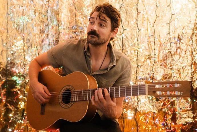 Tim Baker holding his guitar, sitting on a stool in front of many Christmas lights.