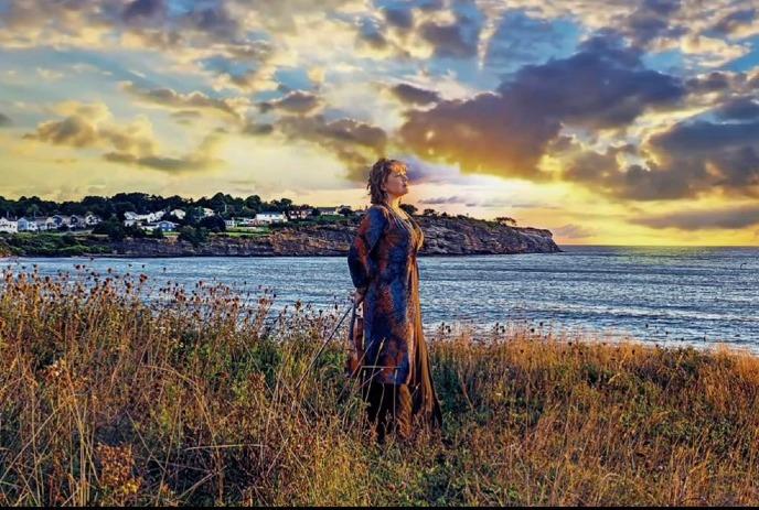 Lucy MacNeil gazes at a sunset on a coastal cliff, violin in hand.