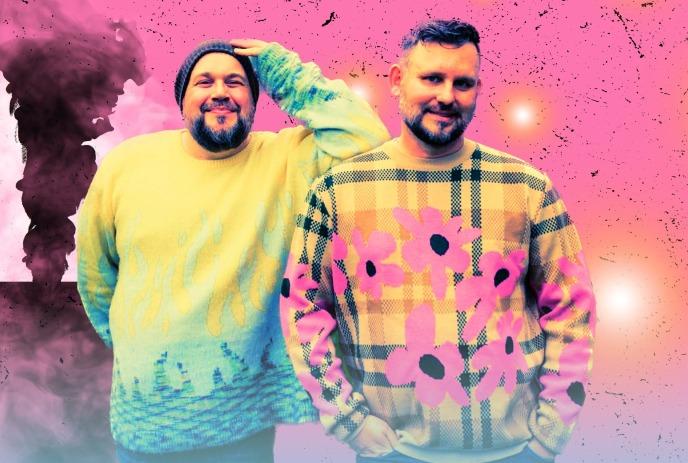 Darcy & Jer pose together against a vibrant pink and smoky background, wearing colorful sweaters.