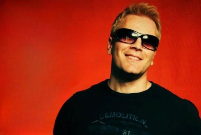 An artist in a black t shirt and black glasses smiling for the picture under a red background.