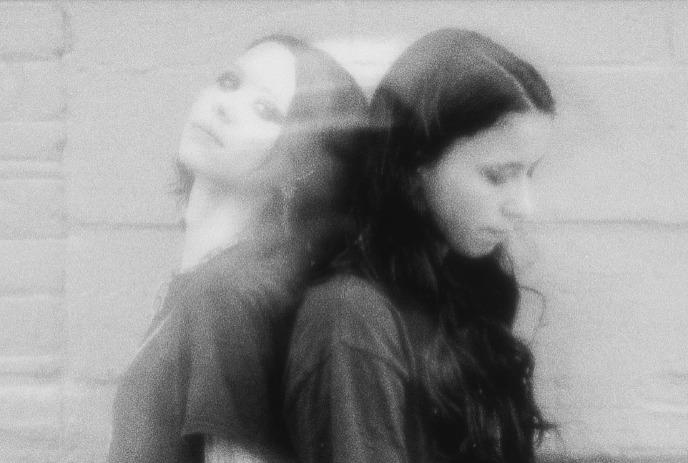 Two female members of the band Softcult stand back-to-back against a brick wall in a grainy black-and-white photo.