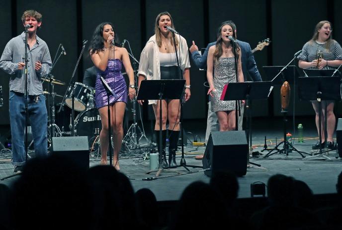 Performers sing on stage with microphones, a drum set, under bright performance lighting.