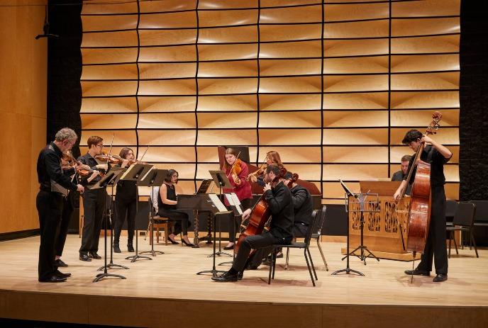 Musicians in formal attire performing on stage with string instruments under a wooden and black background.