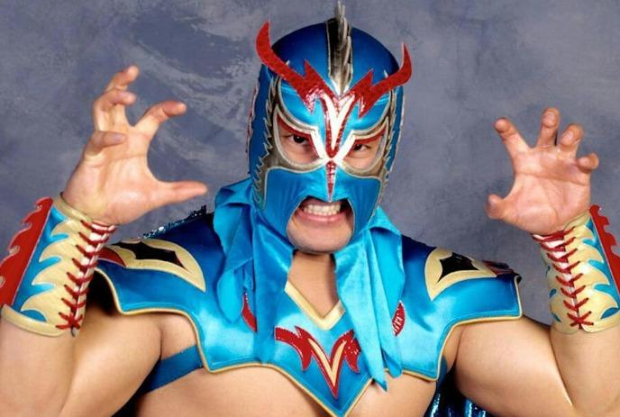 Ultimo Dragon strikes a dramatic pose with raised hands in his colorful blue and red costume.