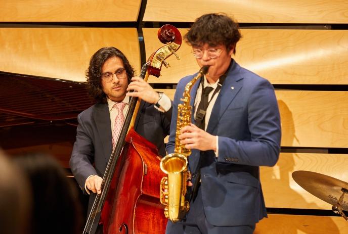 Two musicians perform live, one playing a double bass, the other a saxophone under a wooden background.