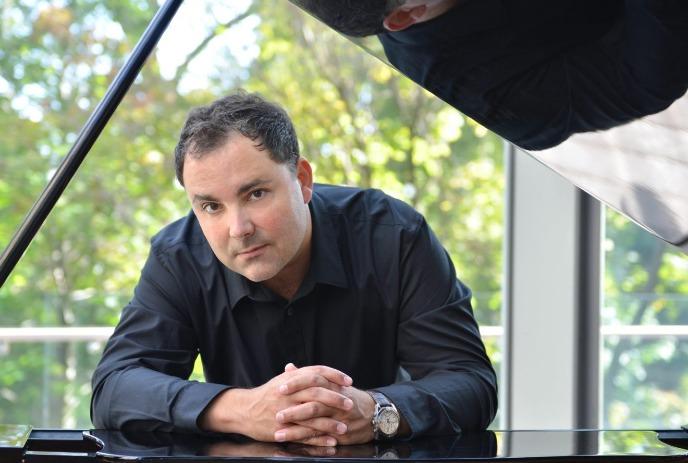 A person in a black shirt with a piano, with greenery visible in the background.