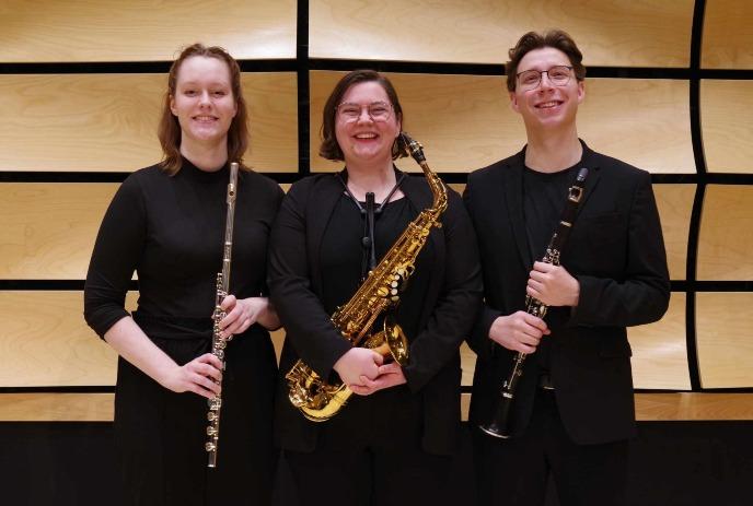 Three females in black attire, holding flute, saxophone, and clarinet, pose before a wooden, grid-like panel.