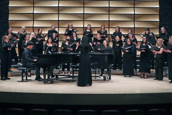 A choir of women in black dresses sings on stage, accompanied by a pianist, with a conductor leading.