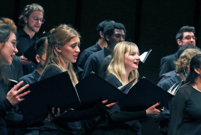 A choir of people, dressed in dark attire, stands holding open books and engaged in a musical performance.