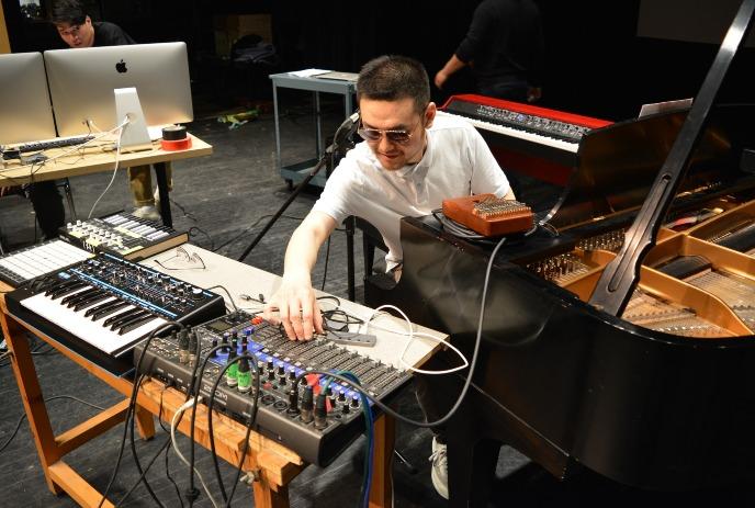 A person seated at a music production setup with keyboards, mixer, computer, and an open grand piano nearby.