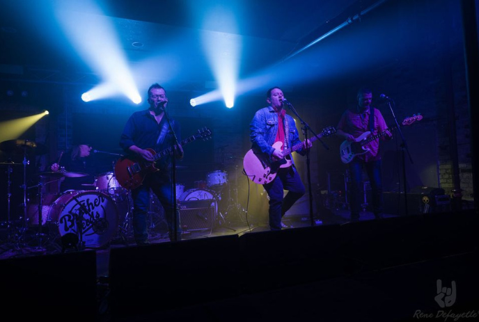 A band performs on stage under blue lights, featuring a drummer, bassist, and two guitarists.