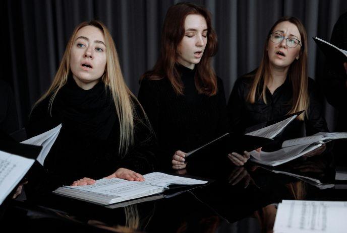 A group of singers holding music sheets. They are in dark clothing, seated at a reflective surface.