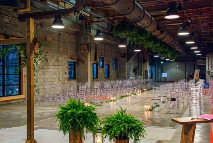 An industrial-style wedding setup at the 'I Do Immersive Wedding Expo' with clear chairs and greenery.