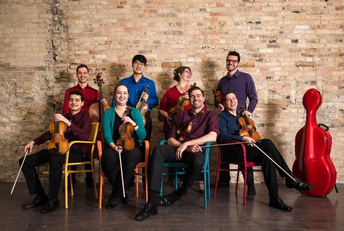 Eight musicians with string instruments, likely a chamber ensemble, in an indoor space with a rustic brick wall.