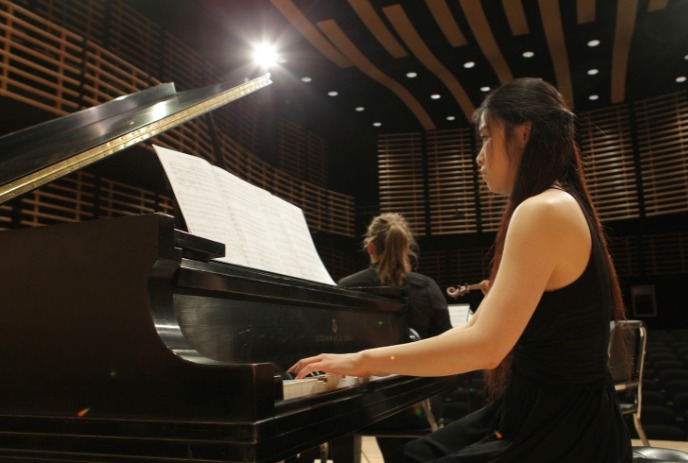 A woman playing a grand piano on stage with sheet music, another musician in the background on a stage.