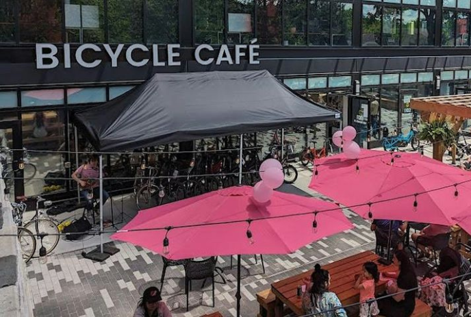 Outdoor café named Bicycle Cafe with pink umbrellas, patrons at tables, and bicycles parked alongside.