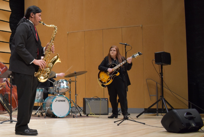 Two musicians on stage, one playing a saxophone and the other an electric guitar, with drums in the background