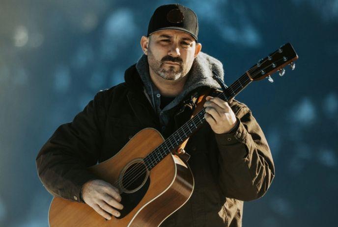 Paul Marier wearing a black baseball cap and jacket, holding an acoustic guitar in front of a blue background.