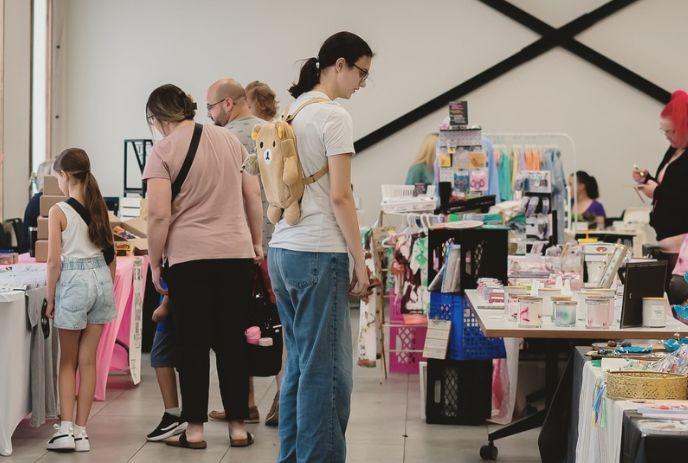 Indoor market with various stalls, shoppers, children, and a person with a backpack shaped like a stuffed animal.