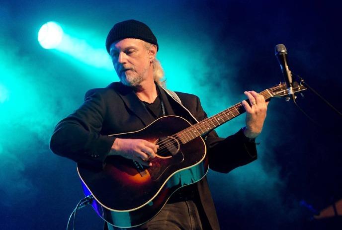 Garnet Rogers playing an acoustic guitar on stage under blue and green stage lights, with a microphone stand nearby.