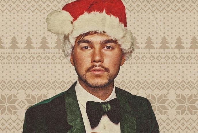 Person in a black suit, white shirt and Santa hat, smiling and standing against a plain background.