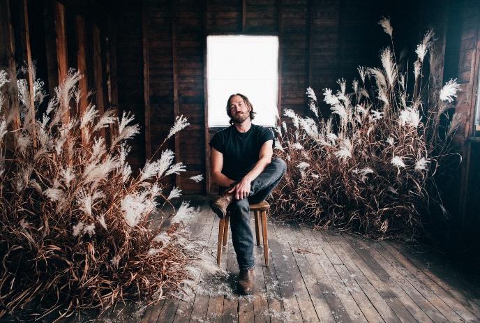 An artist on a wooden stool in a rustic room with wooden floors and walls, decorated with dried plants and a large window.