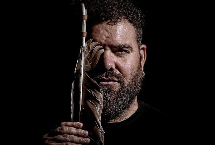 A man with dark curly hair and a beard, holding a large feather that partially obscures one side of his face.