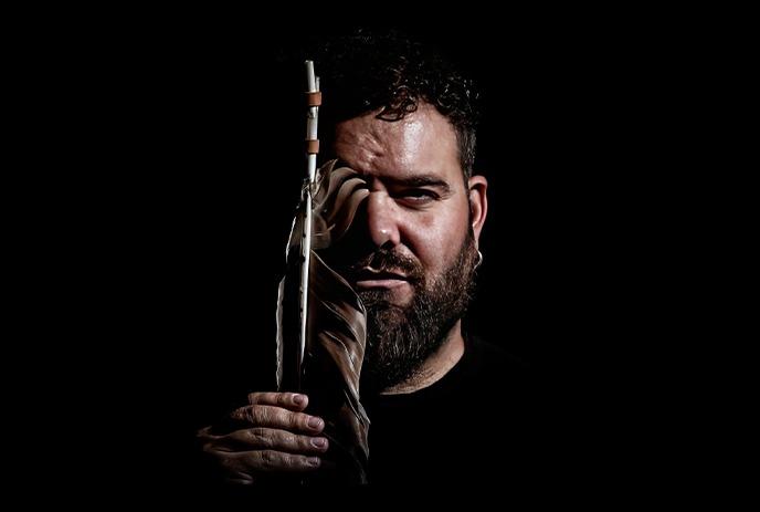 A man with dark curly hair and a beard, holding a large feather that partially obscures one side of his face.