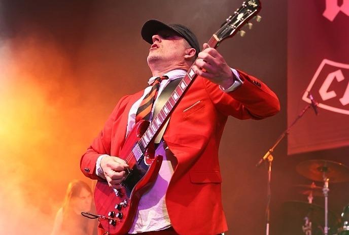 A guitarist in a red suit and tie is performing energetically on stage with a band, with a backdrop featuring the 'AC/DC'.