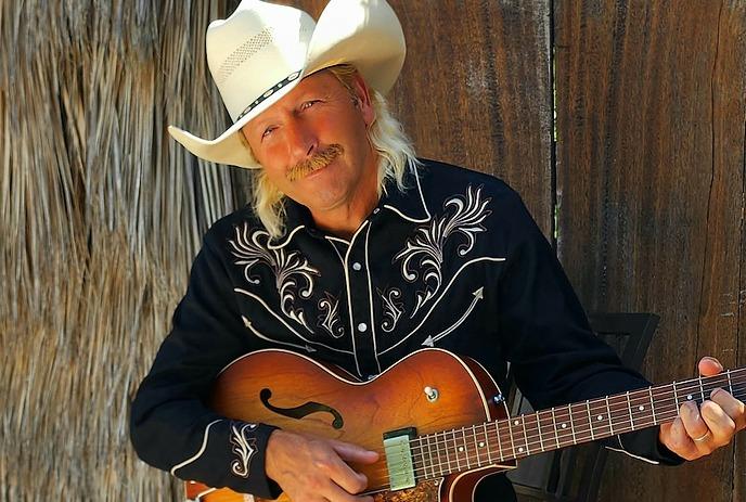 A person wearing a white cowboy hat and a black shirt with intricate white embroidery stands in front of a wooden background