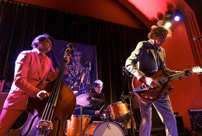 Live music on stage. Three musicians with bass, drums, and guitar under warm red and purple lighting.